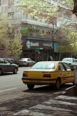 A yellow taxi is parked on the side of a city street, with partially visible license plates. Surrounding the taxi are other parked cars and a building with a shop named 'Benelli'. The trees have budding green leaves, indicating springtime.