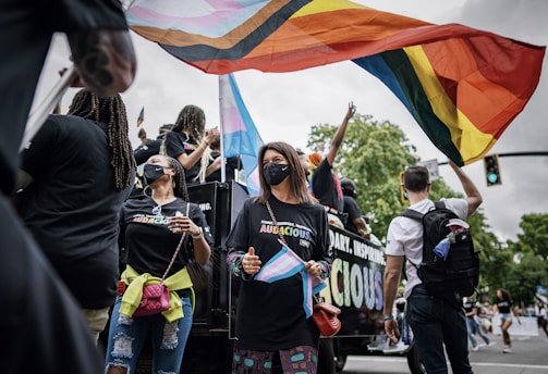 A group of people is participating in a parade, with many holding and waving colorful flags symbolizing LGBTQ+ pride. They wear matching black shirts, some have face masks, and the atmosphere is lively and celebratory. A large truck is decorated with words, and the surroundings feature trees and traffic lights indicating an urban outdoor setting.