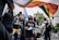 A group of people is participating in a parade, with many holding and waving colorful flags symbolizing LGBTQ+ pride. They wear matching black shirts, some have face masks, and the atmosphere is lively and celebratory. A large truck is decorated with words, and the surroundings feature trees and traffic lights indicating an urban outdoor setting.