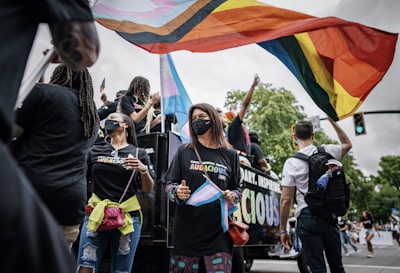 A group of people is participating in a parade, with many holding and waving colorful flags symbolizing LGBTQ+ pride. They wear matching black shirts, some have face masks, and the atmosphere is lively and celebratory. A large truck is decorated with words, and the surroundings feature trees and traffic lights indicating an urban outdoor setting.