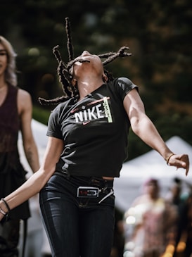 A person with dreadlocks is captured in a dynamic pose, leaning back with arms spread wide, emanating a sense of freedom or joy. Wearing a black 'NIKE' T-shirt and blue jeans, they are in an outdoor setting with blurred people and white tents in the background, creating a lively atmosphere.