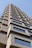 A tall building with a modern architectural design featuring textured concrete and large rectangular windows. The structure rises into a clear blue sky, capturing a perspective that emphasizes its height and geometric patterns.