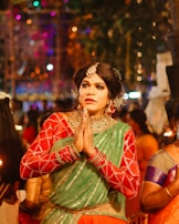 A person dressed in festive traditional attire, with hands in a prayer position. They are wearing a green and red outfit with elaborate jewelry including hair adornments, a nose ring, and a necklace. The background suggests a vibrant and colorful celebration with blurred lights and other people around.