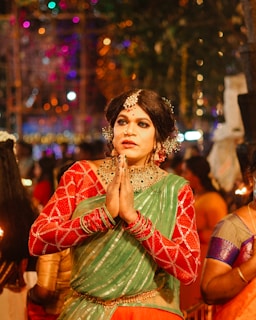 A person dressed in festive traditional attire, with hands in a prayer position. They are wearing a green and red outfit with elaborate jewelry including hair adornments, a nose ring, and a necklace. The background suggests a vibrant and colorful celebration with blurred lights and other people around.