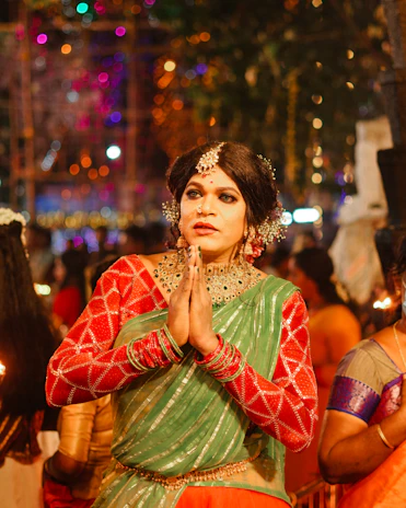 A person dressed in festive traditional attire, with hands in a prayer position. They are wearing a green and red outfit with elaborate jewelry including hair adornments, a nose ring, and a necklace. The background suggests a vibrant and colorful celebration with blurred lights and other people around.