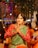 A person dressed in festive traditional attire, with hands in a prayer position. They are wearing a green and red outfit with elaborate jewelry including hair adornments, a nose ring, and a necklace. The background suggests a vibrant and colorful celebration with blurred lights and other people around.