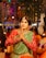 A person dressed in festive traditional attire, with hands in a prayer position. They are wearing a green and red outfit with elaborate jewelry including hair adornments, a nose ring, and a necklace. The background suggests a vibrant and colorful celebration with blurred lights and other people around.