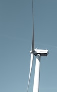 A large wind turbine stands tall against a clear blue sky, with three elongated blades extending from its hub. The turbine's tower is white and cylindrical, supporting the machinery at the top labeled with 'GE Wind Energy 15i'.