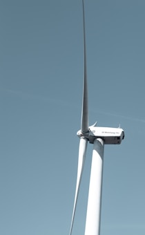 A large wind turbine stands tall against a clear blue sky, with three elongated blades extending from its hub. The turbine's tower is white and cylindrical, supporting the machinery at the top labeled with 'GE Wind Energy 15i'.