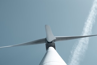 A low-angle view of a wind turbine set against a pale blue sky. The turbine's blades are prominently displayed, emphasizing their size and aerodynamic design. A faint diagonal cloud streaks across the sky in the background.