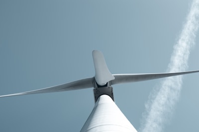 A low-angle view of a wind turbine set against a pale blue sky. The turbine's blades are prominently displayed, emphasizing their size and aerodynamic design. A faint diagonal cloud streaks across the sky in the background.