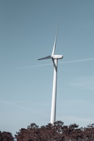 A large wind turbine standing tall against a clear blue sky. It is situated above a forested area with tall trees at the bottom edge of the image. The turbine has a sleek, white structure with three blades. The scene appears calm and serene with a touch of modern technology.