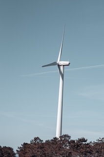 A large wind turbine standing tall against a clear blue sky. It is situated above a forested area with tall trees at the bottom edge of the image. The turbine has a sleek, white structure with three blades. The scene appears calm and serene with a touch of modern technology.