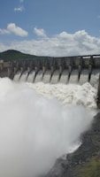 Water rushes powerfully over a series of dam spillways, creating a large, foamy flow. The dam is set against a backdrop of green hills under a cloudy blue sky.