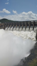 Water rushes powerfully over a series of dam spillways, creating a large, foamy flow. The dam is set against a backdrop of green hills under a cloudy blue sky.