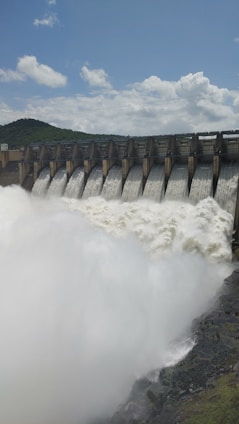 Water rushes powerfully over a series of dam spillways, creating a large, foamy flow. The dam is set against a backdrop of green hills under a cloudy blue sky.