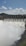 Water rushes powerfully over a series of dam spillways, creating a large, foamy flow. The dam is set against a backdrop of green hills under a cloudy blue sky.