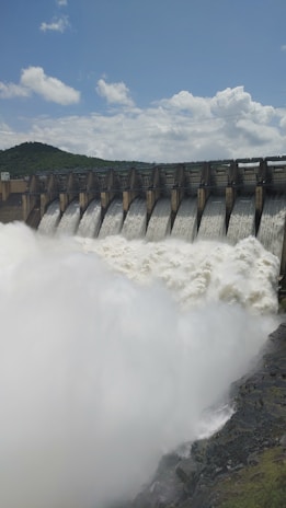 Water rushes powerfully over a series of dam spillways, creating a large, foamy flow. The dam is set against a backdrop of green hills under a cloudy blue sky.