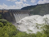 A large dam with a series of spillways releasing vast amounts of water, surrounded by lush green foliage and hills under a partly cloudy sky.