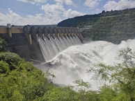A large dam with a series of spillways releasing vast amounts of water, surrounded by lush green foliage and hills under a partly cloudy sky.