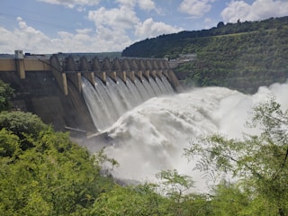 A large dam with a series of spillways releasing vast amounts of water, surrounded by lush green foliage and hills under a partly cloudy sky.