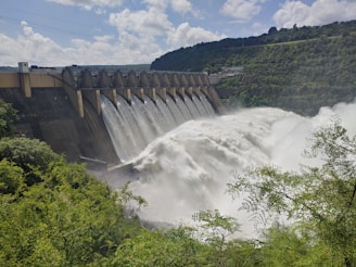 A large dam with a series of spillways releasing vast amounts of water, surrounded by lush green foliage and hills under a partly cloudy sky.