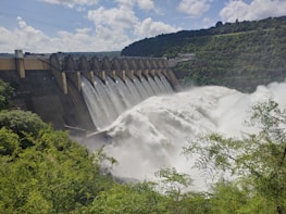 A large dam with a series of spillways releasing vast amounts of water, surrounded by lush green foliage and hills under a partly cloudy sky.