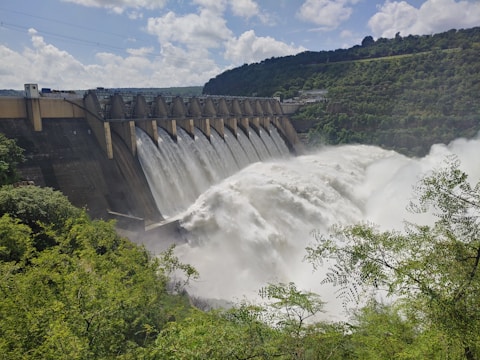 A large dam with a series of spillways releasing vast amounts of water, surrounded by lush green foliage and hills under a partly cloudy sky.