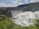 A large dam with a series of spillways releasing vast amounts of water, surrounded by lush green foliage and hills under a partly cloudy sky.