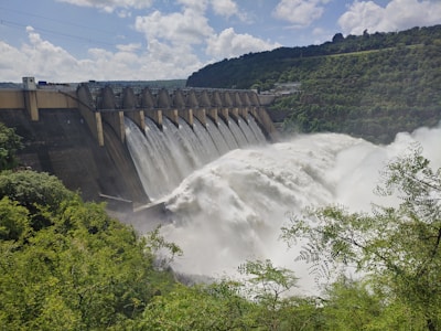 A large dam with a series of spillways releasing vast amounts of water, surrounded by lush green foliage and hills under a partly cloudy sky.