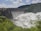 A large dam with a series of spillways releasing vast amounts of water, surrounded by lush green foliage and hills under a partly cloudy sky.