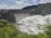A large dam with a series of spillways releasing vast amounts of water, surrounded by lush green foliage and hills under a partly cloudy sky.
