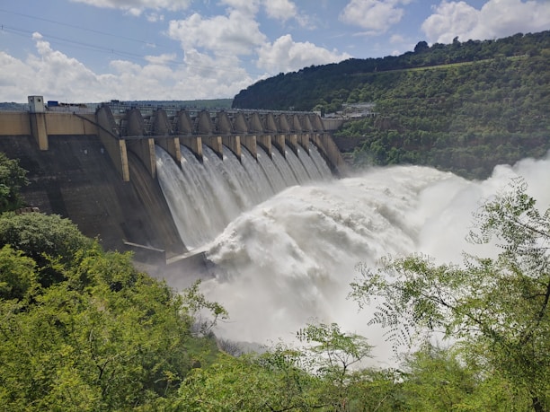 A large dam with a series of spillways releasing vast amounts of water, surrounded by lush green foliage and hills under a partly cloudy sky.