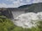 A large dam with a series of spillways releasing vast amounts of water, surrounded by lush green foliage and hills under a partly cloudy sky.