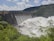 A large dam with a series of spillways releasing vast amounts of water, surrounded by lush green foliage and hills under a partly cloudy sky.