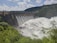 A large dam with a series of spillways releasing vast amounts of water, surrounded by lush green foliage and hills under a partly cloudy sky.
