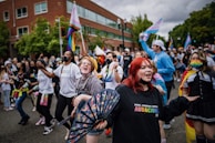 A large, joyous crowd participates in a street parade with rainbow flags and colorful accessories. Several individuals are wearing masks and vibrant outfits, displaying a lively and inclusive atmosphere. People are cheering, dancing, and celebrating together on an overcast day.