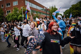 A large, joyous crowd participates in a street parade with rainbow flags and colorful accessories. Several individuals are wearing masks and vibrant outfits, displaying a lively and inclusive atmosphere. People are cheering, dancing, and celebrating together on an overcast day.