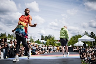 A person is energetically dancing on an outdoor stage wearing a vibrant rainbow-colored fringe top and black leggings, while another person in a green hoodie walks in the opposite direction. An audience surrounds the stage, enjoying the performance under a partly cloudy sky with green trees and white tents in the background.