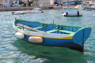 A colorful wooden boat is floating on turquoise water, featuring bright blue and green colors with yellow accents. The boat is equipped with several white fenders. In the background, various other boats are moored near a waterfront with colorful buildings.