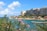 A coastal scene with modern apartment buildings lining the shoreline. There are clear blue skies with scattered clouds and a calm turquoise sea. In the foreground, flowering bushes add vibrant color, while people can be seen enjoying the water near a pier.