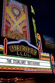 A brightly lit marquee sign with intricate neon lights featuring the Warner Bros logo. The sign advertises 'The Starlight Restaurant' and has art deco style decorations and vibrant colors.