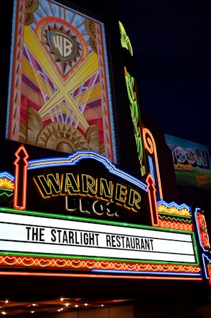 A brightly lit marquee sign with intricate neon lights featuring the Warner Bros logo. The sign advertises 'The Starlight Restaurant' and has art deco style decorations and vibrant colors.