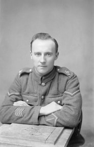 A man in a military uniform sits with his arms crossed on a wooden table. The uniform features buttons and insignia on the shoulders and sleeves.
