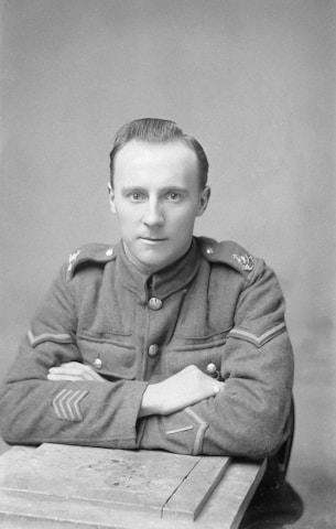 A man in a military uniform sits with his arms crossed on a wooden table. The uniform features buttons and insignia on the shoulders and sleeves.