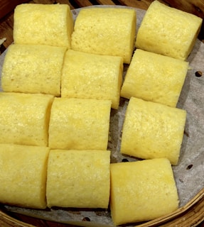 There are twelve cylindrical yellow cake pieces arranged on parchment paper, which is inside a bamboo steamer. The texture appears soft and fluffy.