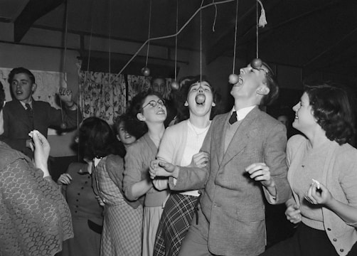 A group of young people engaged in a playful activity at a social gathering. Some are attempting to bite hanging apples attached to strings while others are watching and laughing, dressed in mid-20th-century attire.