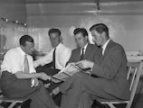 Four men dressed in formal suits are seated in a small, confined room. They appear to be engaged in a serious conversation, with one man holding a newspaper and another pointing at it. The room has a minimalist design, with a few items placed on shelves and tables in the background.