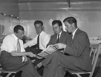 Four men dressed in formal suits are seated in a small, confined room. They appear to be engaged in a serious conversation, with one man holding a newspaper and another pointing at it. The room has a minimalist design, with a few items placed on shelves and tables in the background.