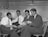 Four men dressed in formal suits are seated in a small, confined room. They appear to be engaged in a serious conversation, with one man holding a newspaper and another pointing at it. The room has a minimalist design, with a few items placed on shelves and tables in the background.